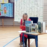 Nauczycielka czyta książkę do mikrofonu przy stoliku z albumami o Podlasiu podczas akcji „Cała szkoła czyta uczniom”..jpg