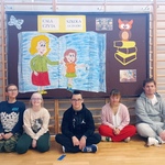 Uczniowie siedzą przed gazetką „Cała szkoła czyta uczniom” z ilustracją otwartej książki i sową na stosie książek..jpg